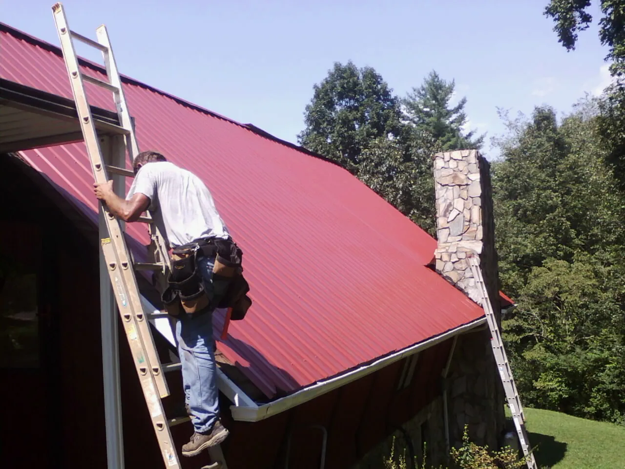 Roofer on a ladder beside a red metal roof and stone chimney — Soffit & Fascia Repair by Roofing Brothers in Starford